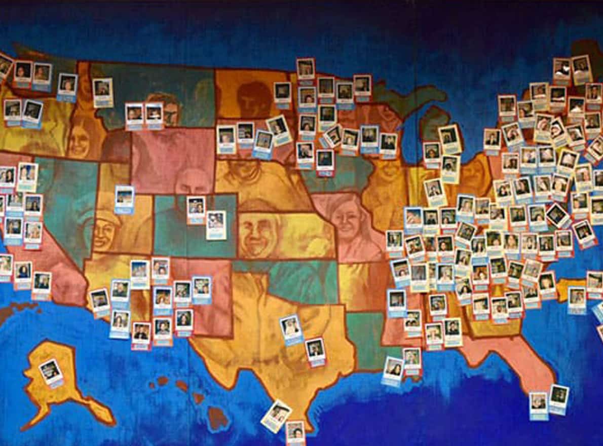 A large painted map of the United States fills a wall, with each state illustrated in warm colors and overlaid with faces of diverse people. Dozens of small instant photo prints are pinned across the map, marking locations throughout the country. The display suggests a community project or event highlighting individuals from different states.