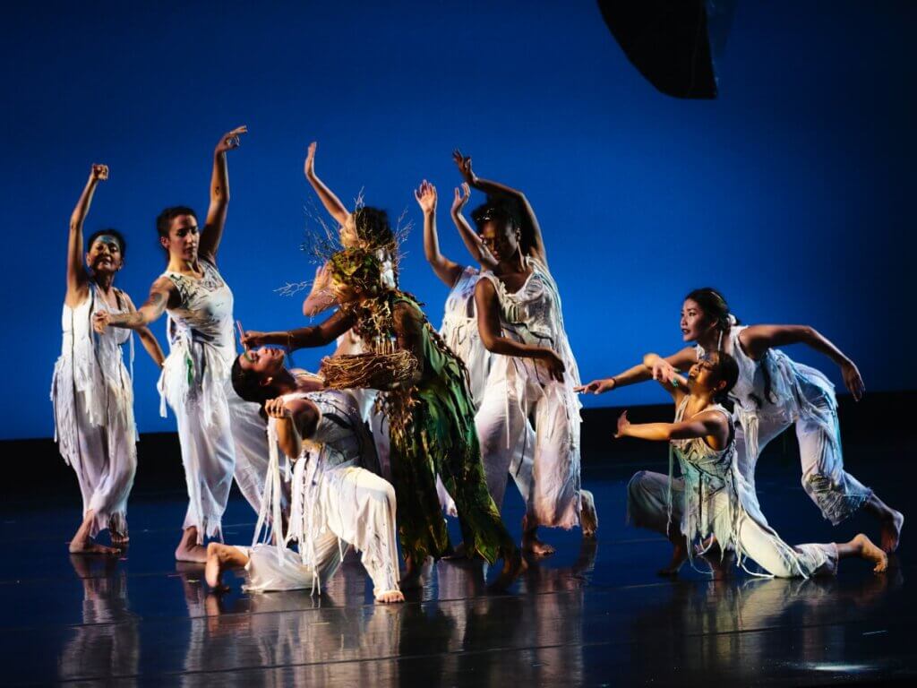 Against a lit, blue background, five femmes in shredded, white costumes stand with open arms, surrounding one woman who kneels in a backbend. A non-binary artist in green wearing a crown of golden twigs paints the kneeling woman’s forehead. To the right, one woman kneels, facing the rest, with arms rounded, her sternum opening to the group. Another woman leans over her and reaches toward the group. All the artists are of various medium-to-dark brown skin tones. 