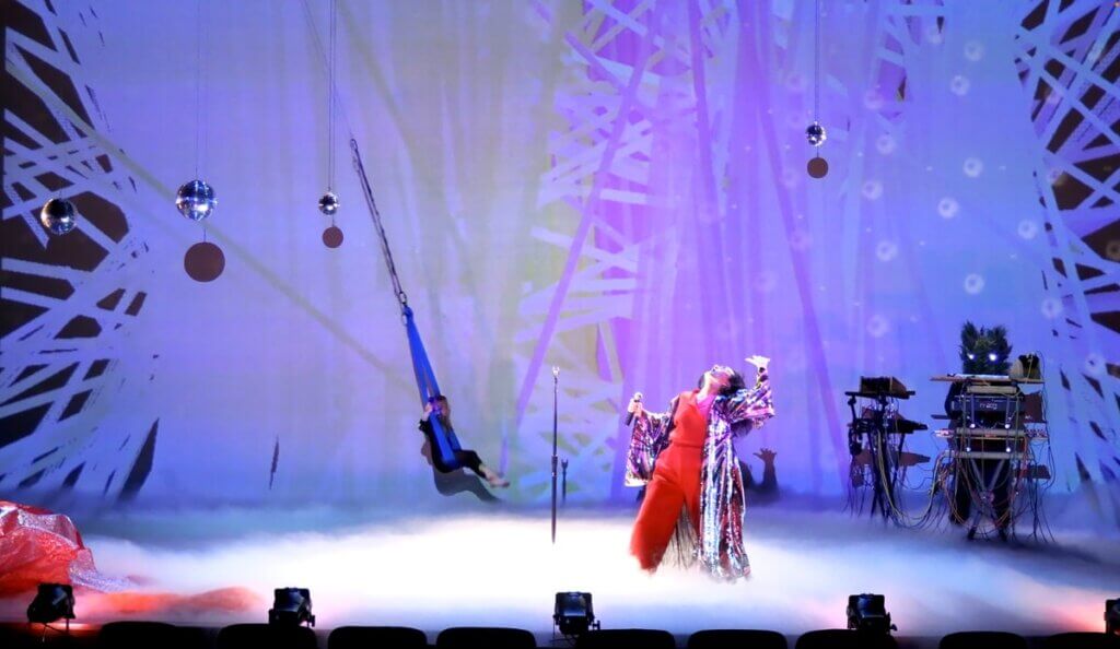 A dark-haired, medium-skinned woman wearing a red jumpsuit and a full-length sequined robe is downstage, holding a mic, her head thrown back, with the mic stand next to her. In the back of the stage, a little girl with blonde hair swings on a blue cloth swing, and a person with a tree branch face and glowing eyes operates a synthesizer and sound machinery. Disco balls hang from the ceiling. Smoke covers the performers' feet. A projection of light beams and lasers takes up the entire back wall of white geometric shapes.