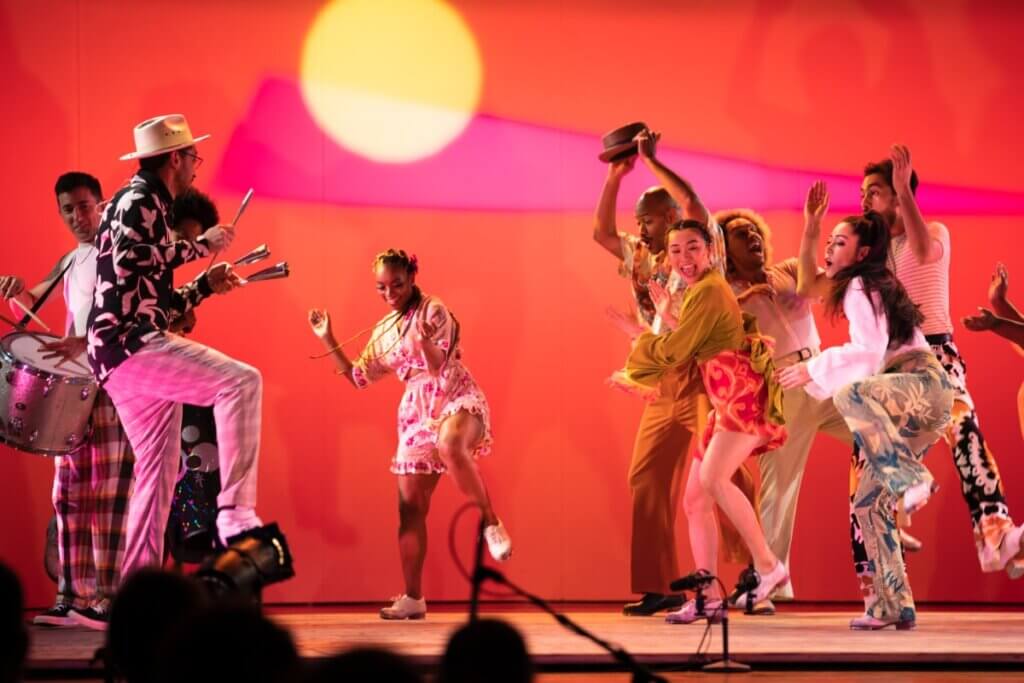 A female dancer with braided hair and wearing a floral-print dress is mid-step, ready to clap her hands and stomp her left foot, smiling as she takes a tap solo in front of a multiracial company of dancers and musicians. All are dressed in colorful clothes, in vibrant orange lighting with a pink triangle and yellow circle in the background, evoking a festive, carnival-like atmosphere.