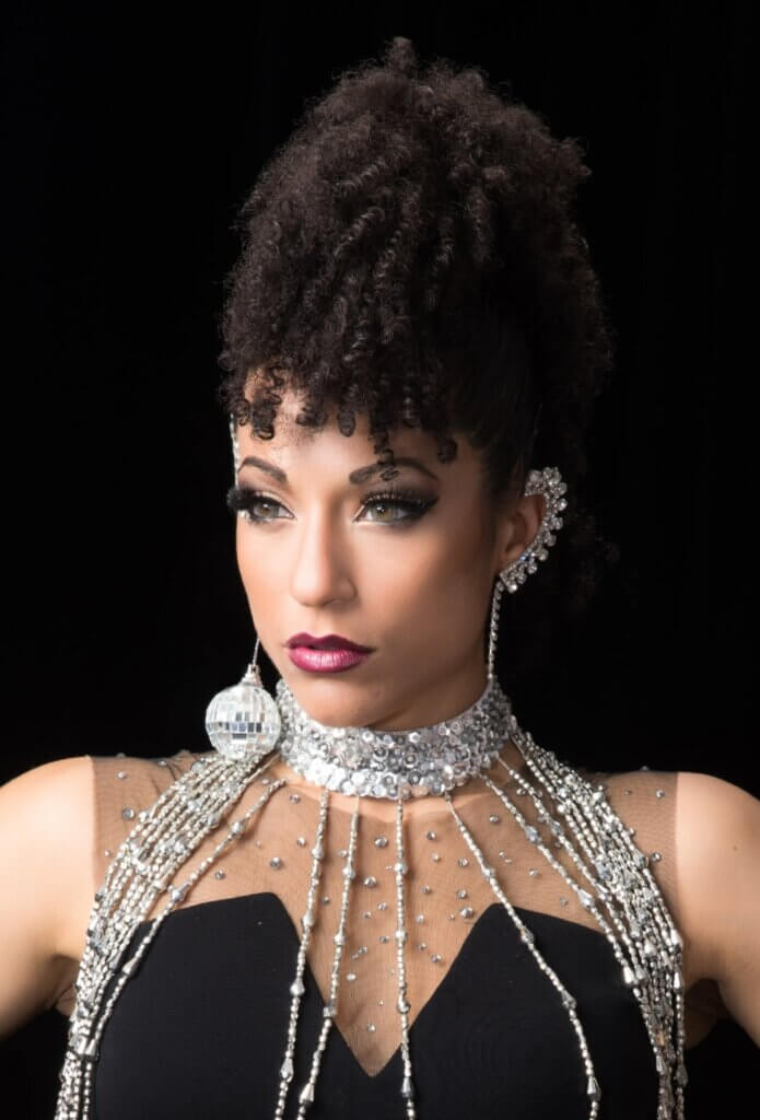 A tan-skinned, multi-racial woman with dark hair and hazel eyes is shown from the shoulders up. She has tight curls pulled up high on her head with bangs and is wearing a black strapless top with rhinestones lining her neck area. She has a silver, sequined choker that drips several strips of silver beads down past her top. She wears a dangling disco ball earring in one ear, and the other ear is lined with silver rhinestones that drip down into a single strand from the ear lobe.