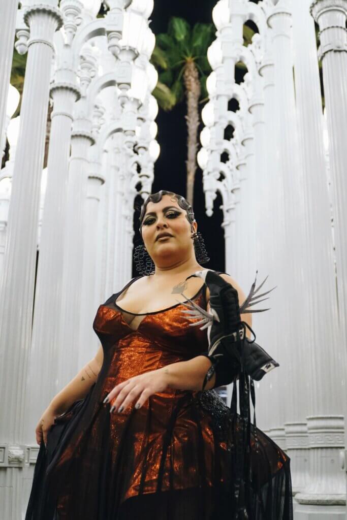 A medium-skinned performer stands tall in front of numerous white columns. They wear a dark amber dress fitted with a corset top and black mesh detailing at the shoulders and from the waist down. On the performer’s left arm, they showcase a bird puppet with silver feathers, a black body, a white head, and a black beak with ribbons descending. Their nails are black, and their hair is in an updo with waves, gold and black eye makeup, and large black earrings.