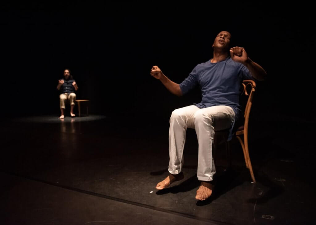 In the foreground, a Black man with a soft blue shirt and white pants sits with his eyes closed and head tilted back in the midst of a song or a shout. His feet are lifted off the floor. In the background, a white-skinned person with dark hair and a dark beard is seated, also calling or speaking, though their image is slightly blurred. The background figure also has one foot slightly lifted and hands raised. Each figure is lit from above and separated by pools of darkness.