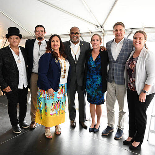 This image shows a group of seven people standing together under a tent, posing for a photo. They are dressed in a mix of formal and semi-formal attire, with some individuals wearing distinctive cultural or traditional clothing. They appear to be at an event or gathering, and everyone is smiling warmly at the camera.