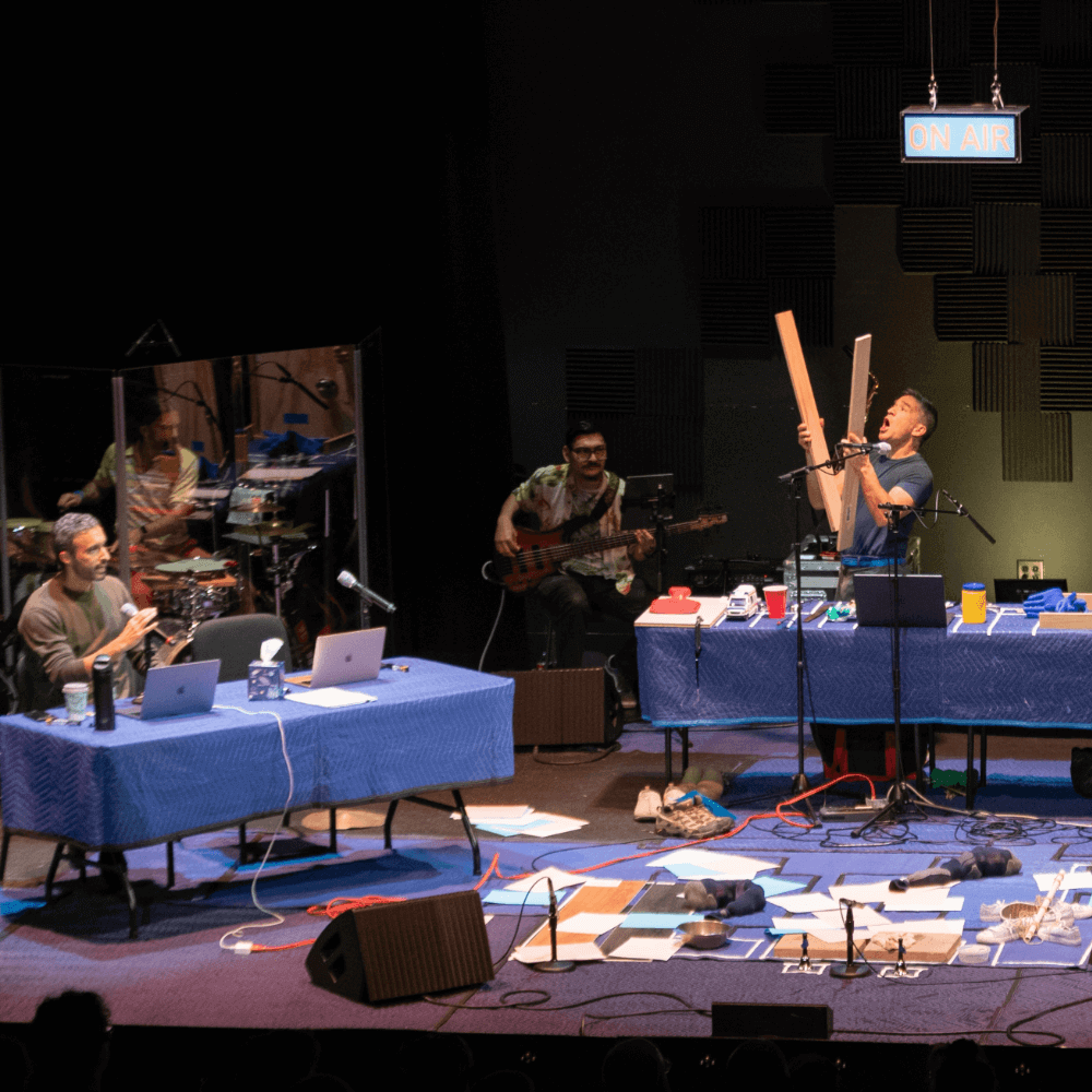 Four performers on a stage strewn with cords, papers, and bags. There are two long tables covered in blue plastic. At the table on the left, a man holding a microphone sits behind two open laptops. At the table on the right, a man is standing and holding two long slats of wood in a way that suggests he is banging them together. In the background, a drummer and a guitarist are performing.