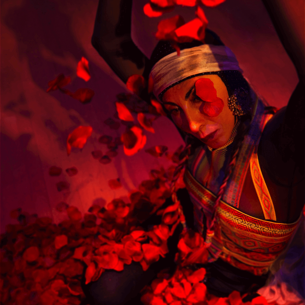 Overhead performance shot of Nejla Yatkin bathed in red light and sitting with her legs folded under her in a pile of rose petals. She is wearing a colorful patterned garment and a headscarf, and holding more rose petals above her head so that they fall gently onto her face. Two large petals rest on her cheek and partially obscure one eye.