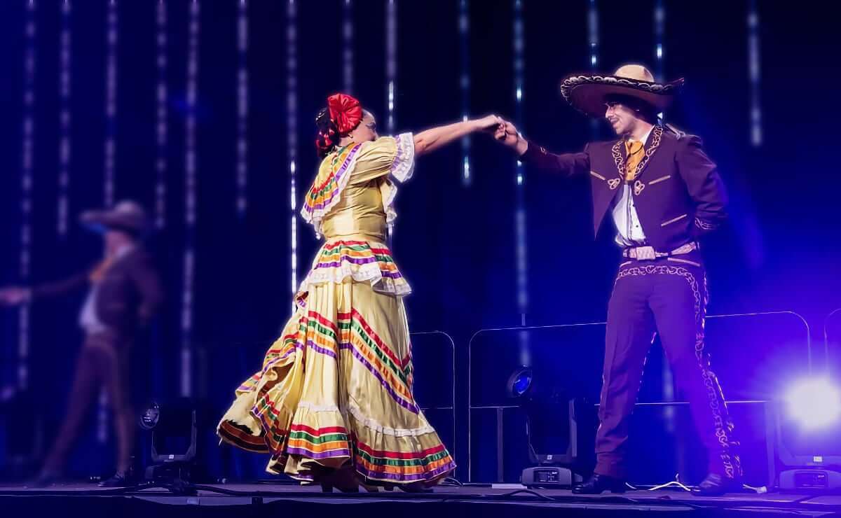 Two performers in traditional Mexican attire dancing on a blue-lit stage under bright lights. The woman is wearing a red headscarf and a ruffled yellow dress with red, green, orange and purple stripe accents. The man wears a black or dark blue charro suit with intricate embroidery and a wide-brimmed sombrero. The background features blurred lights and another performer in the distance.