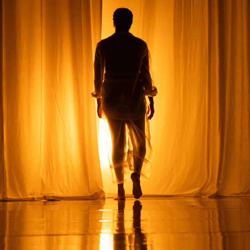 A barefoot person in silhouette walks away from the camera toward a bright golden light behind tall flowing curtains.