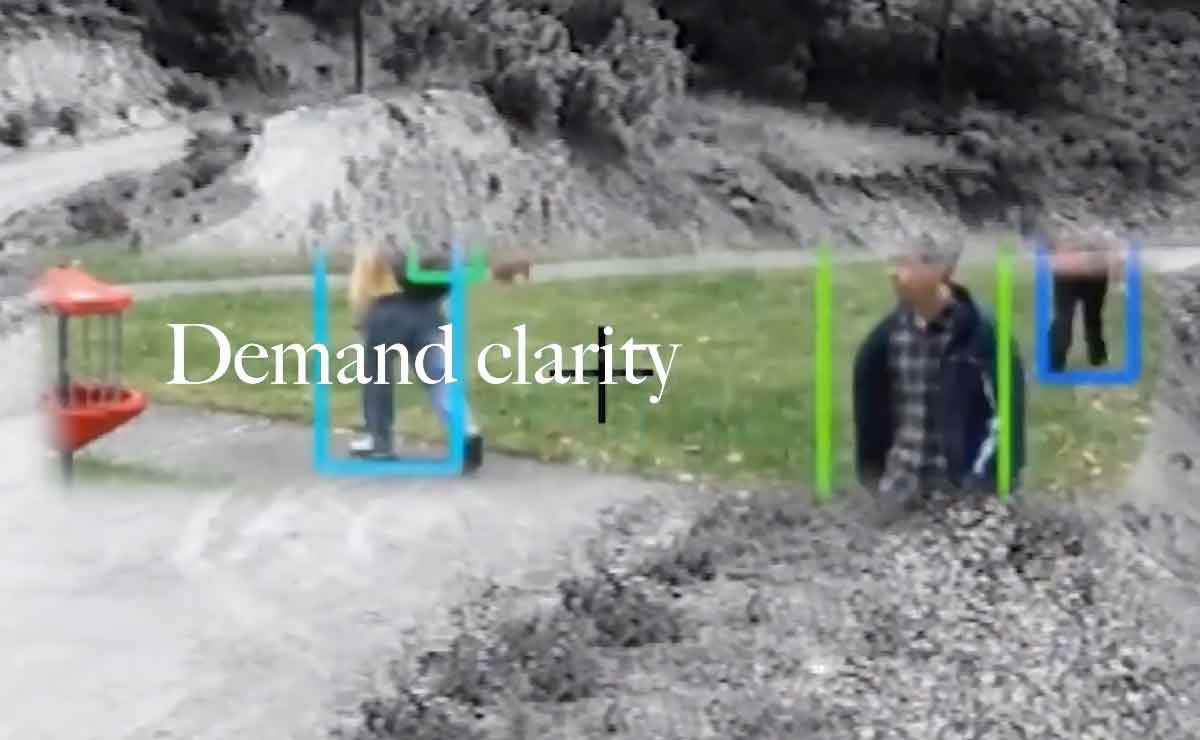 A frame from a video essay. It combines footage from two different preexisting pieces of footage. A cropped rectangular section of color footage with soft edges floats over a black and white image of a dirt road that curves through low hills, The color footage shows a closely cropped strip of park with green grass and concrete footpaths. Three people are walking separately across the park. Around each person is a rectangular outline, signifying that they are being identified and followed with intelligent tracking software and security cameras.