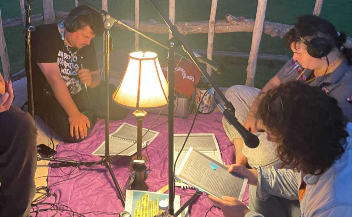 Three actors perform a dramatic reading at dawn at Nanih Bvlbancha. The actors are sitting on blankets with a tall shaded lamp between them. They are looking down at their scripts. Behind them is an irregular fence made out of rough-hewn branches and thin logs.