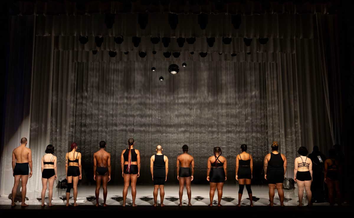 A row of dancers in black shorts, some shirtless and some in sleeveless tops, stand in a row at the front edge of a large, empty stage with their backs turned to the audience. At the back of the stage is a blank gray curtain.