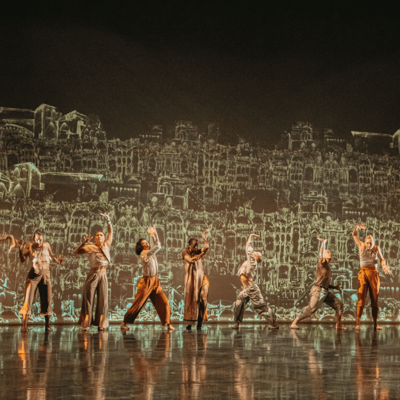 A line of seven dancers of various brown skin tones, wearing loose fitting clothes in muted shades of brown, gray, orange, and yellow, stand in various poses on a reflective stage, against a 15-foot-tall backdrop of overlapping hand-drawn sketches of buildings and arches in light green against brown. The image is cropped to suggest that there are similar dancers, not in this photo, extending to either side on the stage.