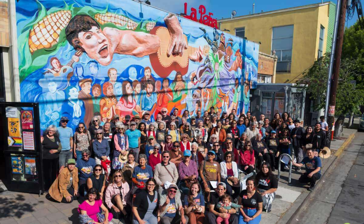 Group photo of staff and friends of La Peña posing together outside, in front of a colorful mural that includes images of corn, water, a singer, guitarists and other musicians,and people watching and celebrating. On the roof is a sign that reads “La Peña” in large red letters.