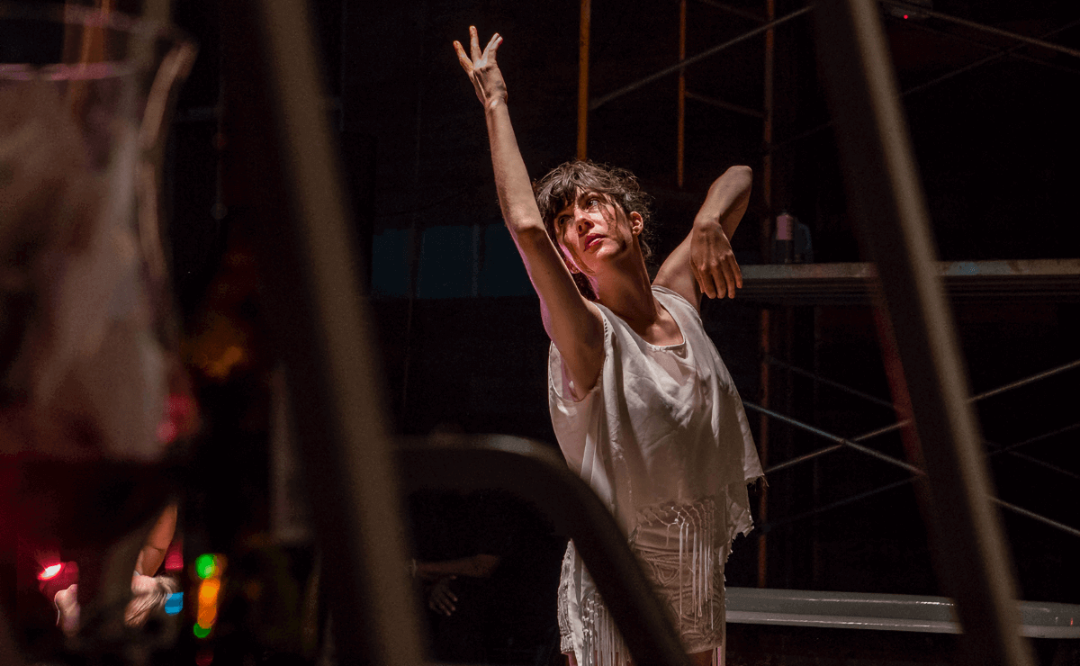 A female dancer in tattered white clothing stands in a pool of light, surrounded by darkness and irregularly placed scaffolding, and extends her right arm above her head as she looks up into the distance.
