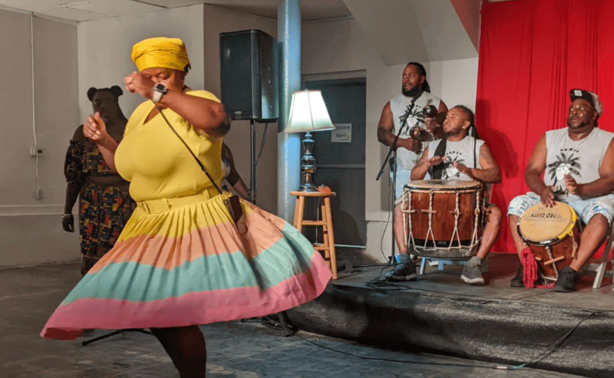 A Caribbean woman wearing a yellow head wrap and a matching yellow dress with wide orange, blue, and pink stripes on the bottom dances in front of a small stage. On the stage, two seated men play drums while a third man stands behind them shaking maracas.