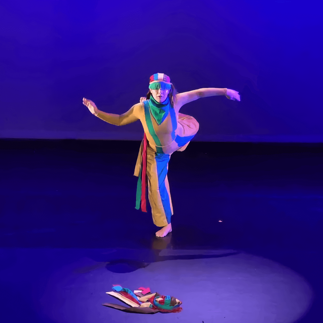 On an empty stage lit entirely in blue, a lone female dancer stands on one bare foot and holds the other extended behind her, with her arms stretched out for balance. She is wearing a loose-fitting yellow pant with a wide blue vertical stripe down each leg, and a matching yellow long-sleeved top with a green vertical stripe running down the right side of her torso like a suspender, and a matching blue vertical stripe running down the left side. A green kerchief is tied around her neck and she is wearing a colorful striped hat with green fringe hanging over her eyes. Before her on the stage floor is a colorful, discarded costume piece of unknown function.