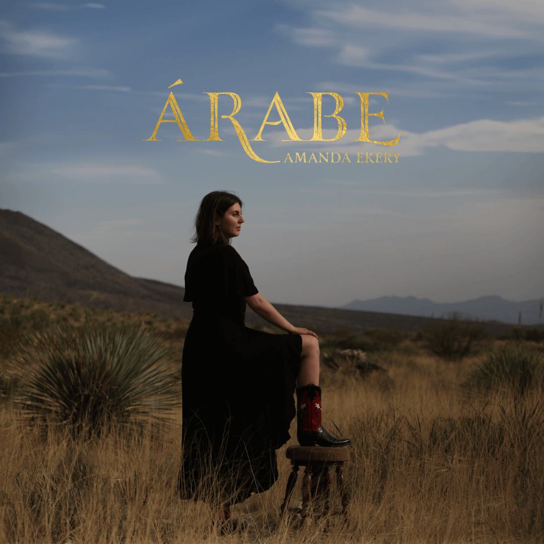 Amanda Ekery poses in profile among the foothills of the southwestern United States. She is wearing a long, plain black dress with sleeves that come to the elbows, and red cowboy boots. She has one foot lifted and placed on a short stool. A pale blue sky, softened by gentle clouds that turn to a white haze in the distance, fills the upper half of the image. Printed over the sky in gold script is the title “Árabe” in all caps. In smaller text below that is the name “Amanda Ekery.”
