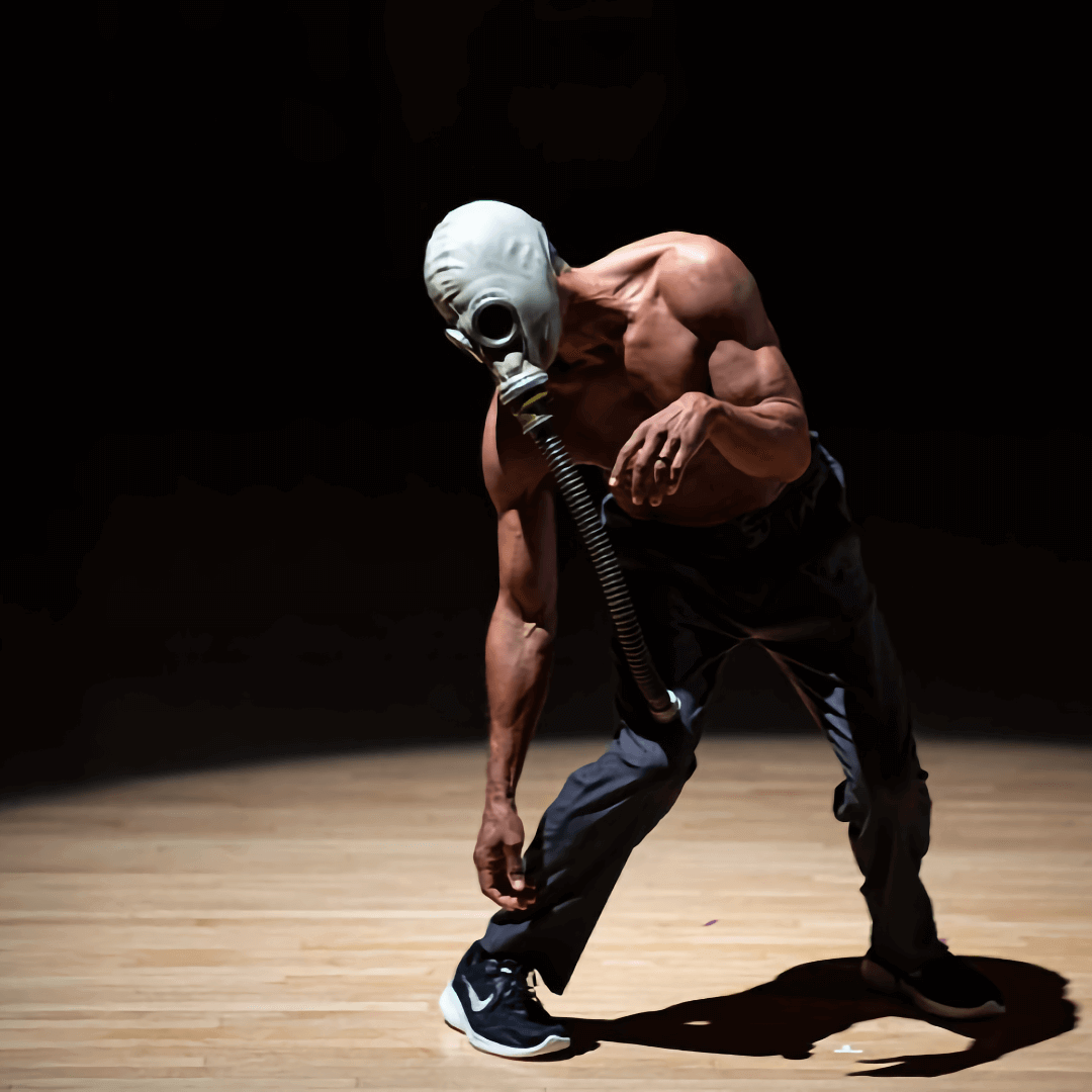 A man in a stark pool of light leans heavily to the left, with one arm dangling low to the ground. He is wearing an old gas mask, and the breathing tube also dangles from his face, disconnected from any source. His upper body is bare, revealing his medium brown skin and the musculature of his torso; on his lower body, he wears dark pants and black running shoes with white swooshes.