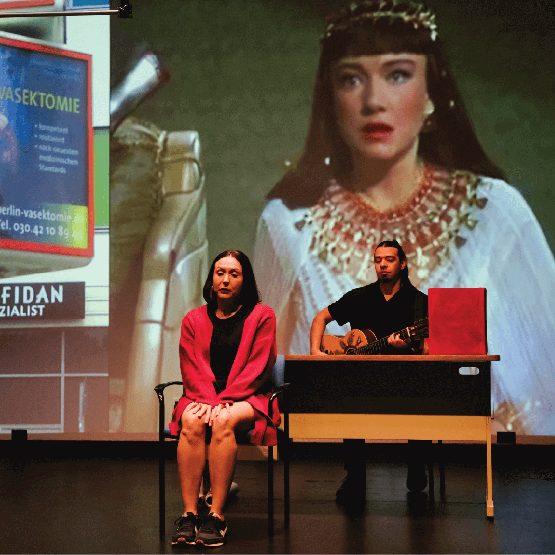On stage, a woman in a short black dress and a long red coat sits with her hands drawn together and pressed between her knees, while a man in a short-sleeved black shirt sits at a desk behind her and plays guitar. Projected onto the back of the stage as if on a movie screen is a 15-to-20 foot tall image of a young woman wearing a Hollywood-style “Egyptian” costume.