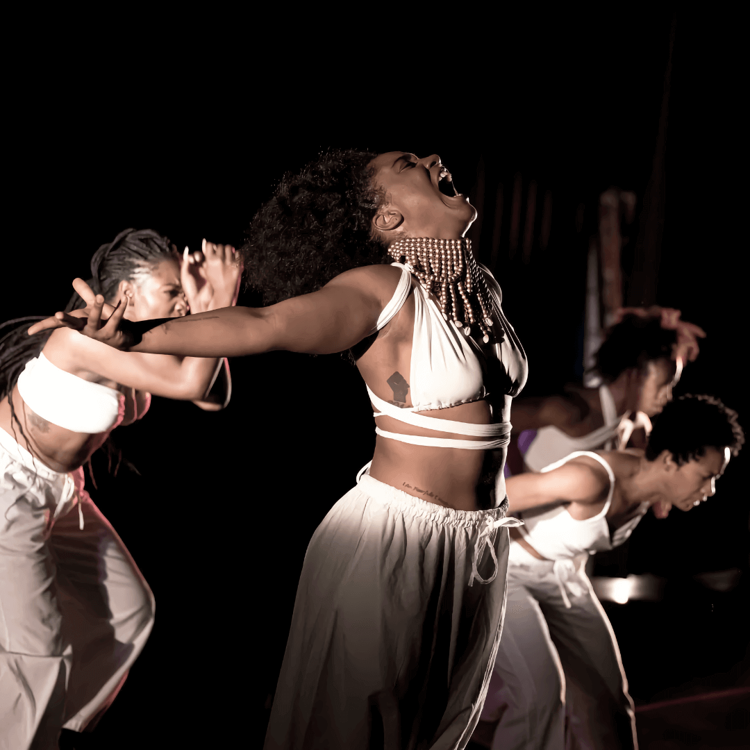 Four Black female dancers, wearing loose white pants and white tops, perform in a spotlight on a dark stage. The dancer in the foreground has her arms and head thrown back as she cries out. Each of the other three dancers behind her bend forward at different angles; one looks directly ahead, her arms held in front of her face, while the other two look down at the ground while throwing their arms back.