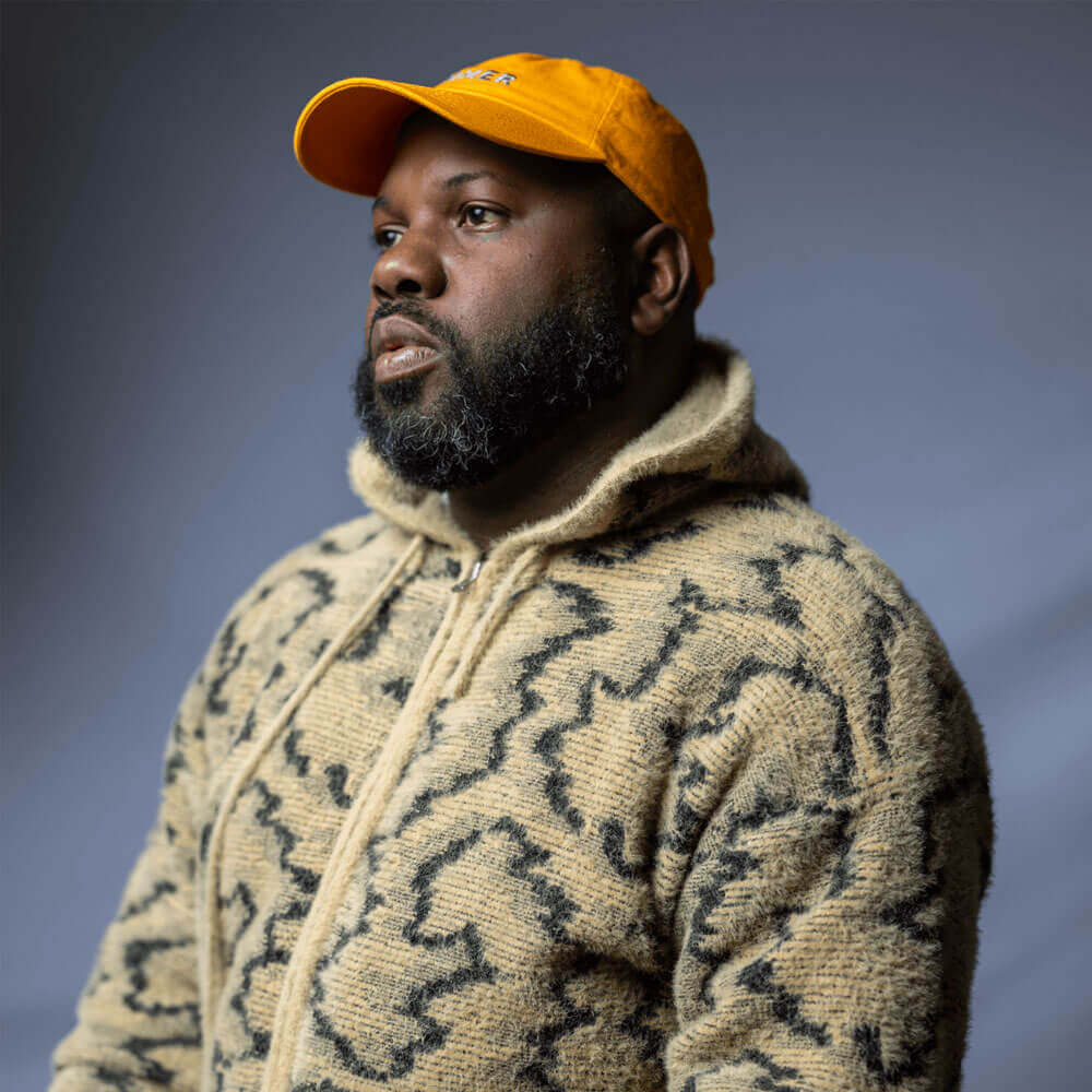 A dark-skin Black man, in a black and tan hoodie and an orange cap. It is a 2024 self-portrait of L. Kasimu Harris, a writer and artist.