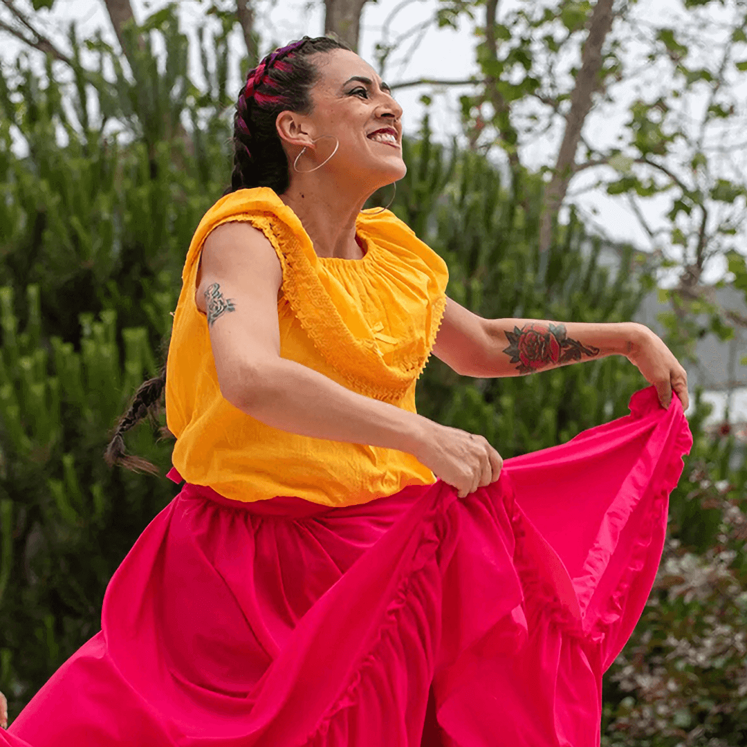 A smiling woman with black hair woven into two long braids with crimson highlights dances in an outdoor setting. She is wearing a loose-fitting marigold-colored sleeveless blouse and is holding the front of her flowing, bright red skirt as she dances.