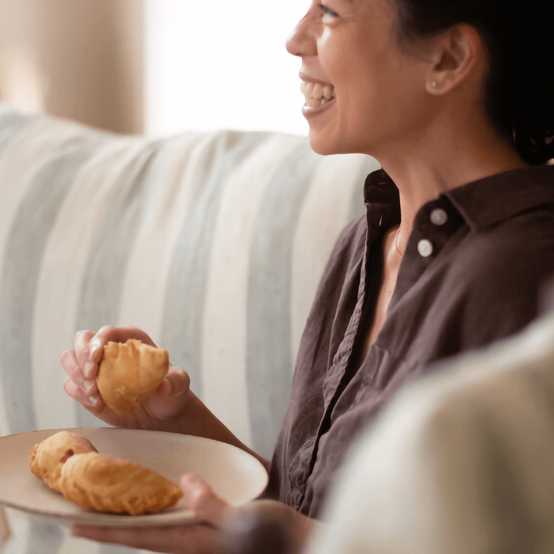 The artist Mary Prescott, wearing a chocolate brown button-down shirt, sits in profile in a brightly lit room decorated in neutral colors — ivory, pale blue, light brown — and smiles as she holds a plate of what appears to be empanadas or pastries.