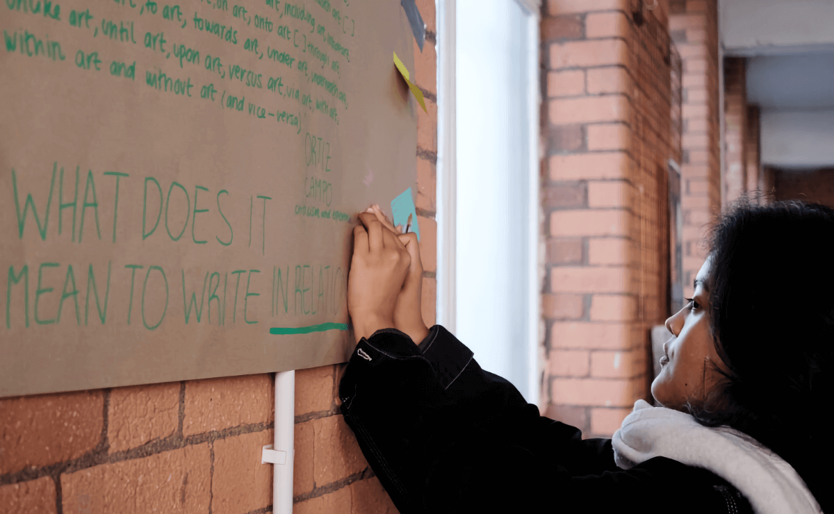 A woman with dark hair affixes a sticky note to a large sheet of brown butcher paper taped to a brick wall. The paper is covered in green lettering, most of which is illegible, but in the foreground is the question fragment, “What does it mean to write in relation…”