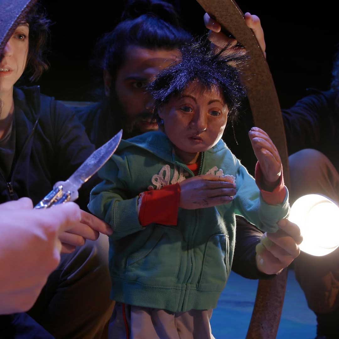 Film still of a refugee puppet with dark hair and a green jacket holding his hands in front of his face. Two puppeteers are visible behind him, holding hm in position. A third puppeteer is just out of frame on the right, holding a bright light pointed at the puppet. In the foreground on the left, a fourth unseen puppeteer’s hand extends into frame, holding an open butterfly knife with the blade pointed threateningly at the puppet’s face.