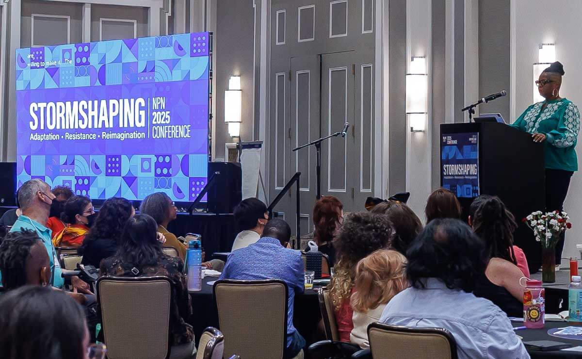 A large,ballroom-style conference space with a seated audience facing away from the camera and looking to the right side of the image. On the right, a Black woman stands behind a podium and speaks to the audience. On the left side of the image, in the corner of the room, a large digital screen faces the audience and displays the blue interlocking geometric shapes of the conference branding, with the words “Stormshaping, NPN 2025 Conference” in large white letters.