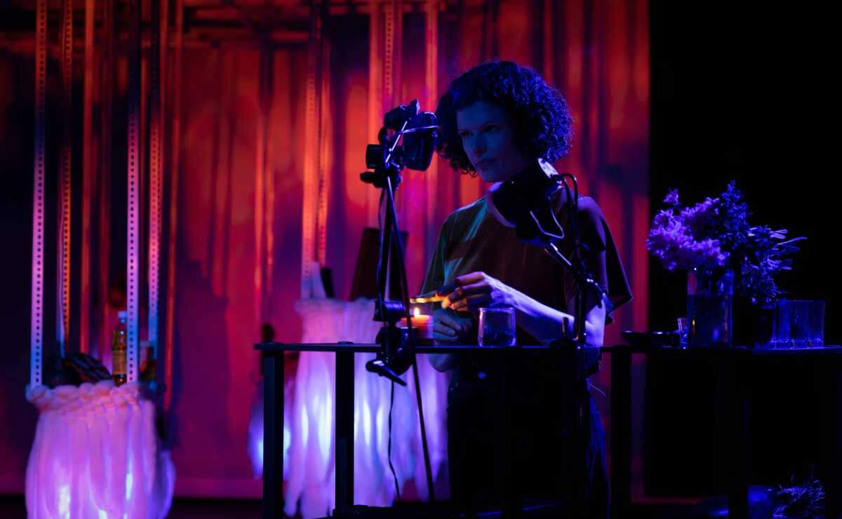 A darkened performance space lit from the rear with red and blue lights. In the background is a stage curtain. In the foreground, backlit and almost entirely in a deep blue shadow, a woman with full, wavy hair that reaches her neck stands at a simple table. She is wearing a short-sleeved dress and her bare forearms rest against the edge of the table. She appears to be holding an unidentified object. Clamped to the table in front of her and to her left are two large lamps on adjustable armatures. 