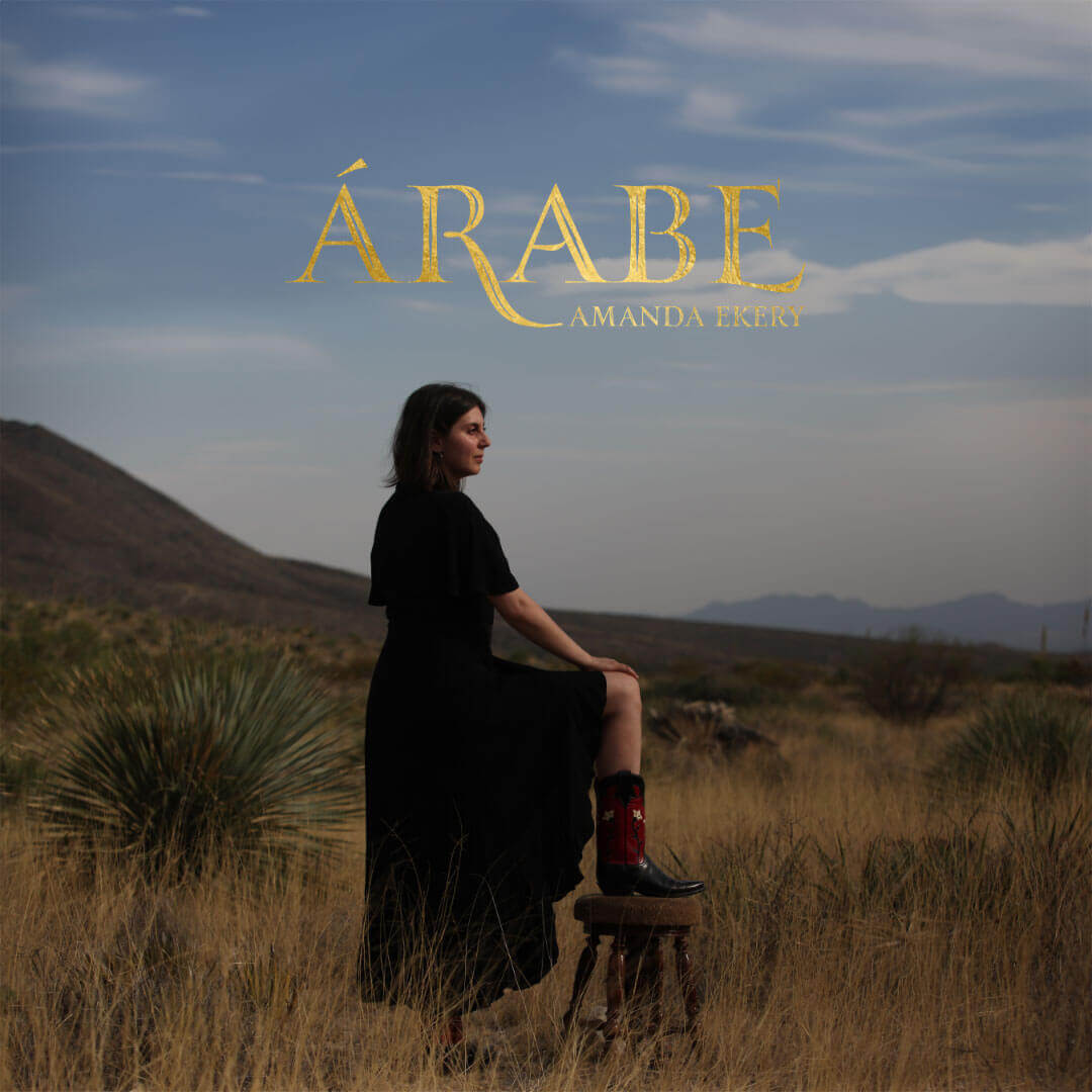 Album cover for Árabe by Amanda Ekery. An exterior photo of the dry scrub brush and low hills of the southwestern United States. The artist Amanda Ekery, a light skinned woman with shoulder length dark hair, stands in profile in the center of the image with one foot lifted and placed on a stool. She is wearing a long black dress with sleeves that stop just above the elbow, and dark red and black traditional cowboy boots. The knee and upper calf of her raised leg is visible, and she has a hand resting on her knee.