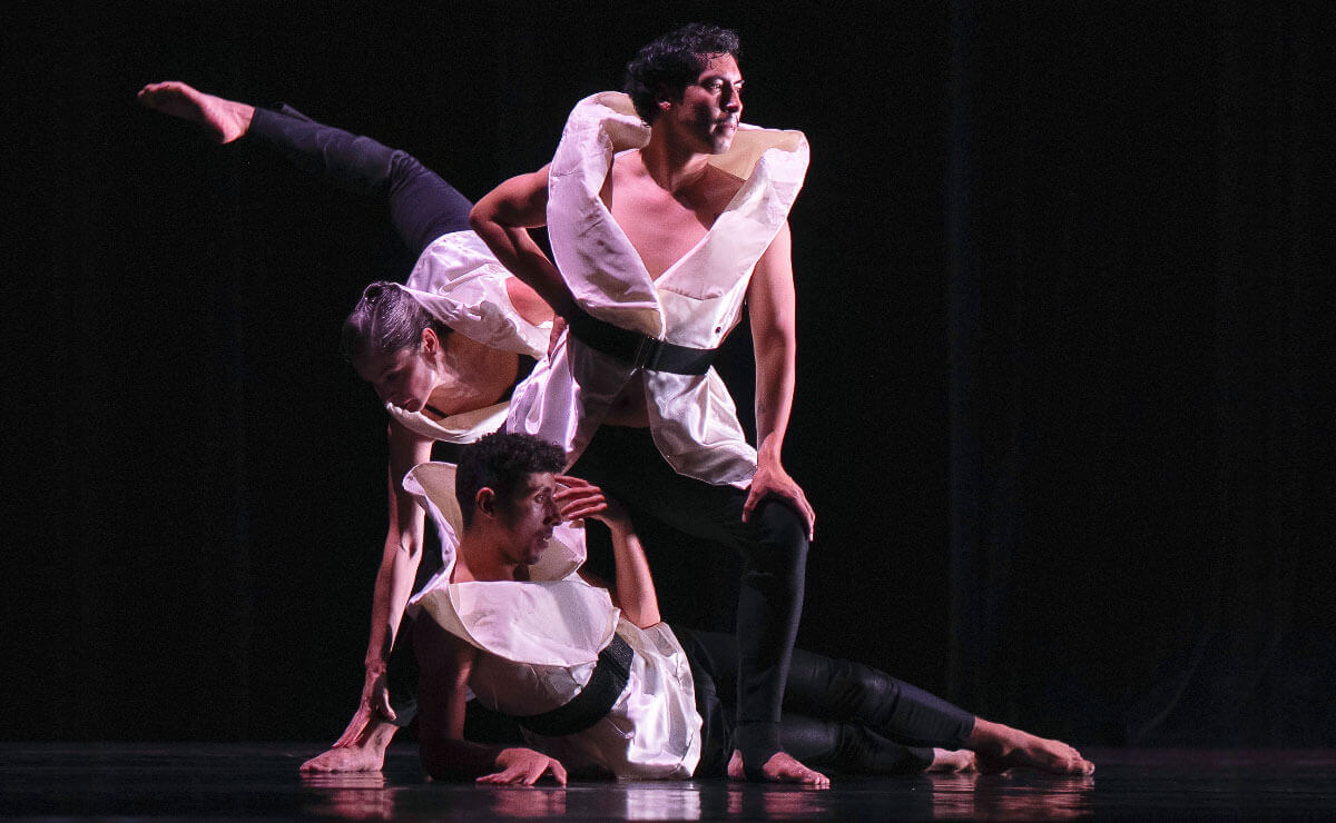 Three barefoot dancers wearing matching costumes of large white jackets with oversized collars, wide black belts, and black leggings, pose on a darkened stage in a scene from The Dulling Effect. One dancer is reclined on the stage and supporting his raised upper body with his right forearm while touching his forehead with his free hand. A second dancer is standing astride the first and leaning forward slightly, one hand on his hip and the other on his knee, as he stares confidently into the distance. The third dancer is behind the other two, extending a hand down one leg while extending the other leg into the air and over her head. Taken together, the trio create a cluster of white fabric and brown skin in the center of the stage, with a gently curving line passing diagonally through the cluster formed by the extended leg in the upper left and the reclining dancer's legs in the lower right part of the frame. 