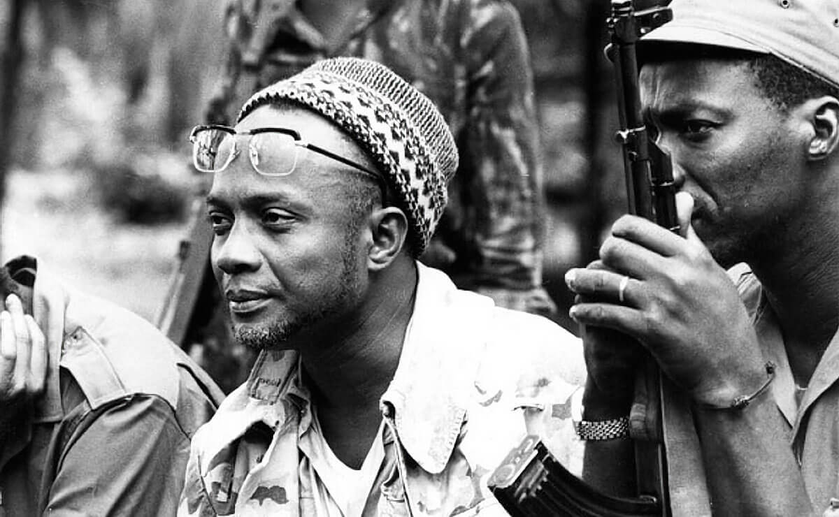 Black-and-white photograph of Amílcar Cabral, wearing a patterned knit cap with glasses perched on his forehead. He is dressed in a camouflage jacket over a light shirt and has a calm, thoughtful expression. To his left is another man who is holding a rifle, suggesting a military setting.