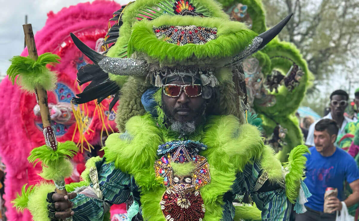A Black man with a gray and black beard and orange-tinted sunglasses poses for the camera during an outdoor festival. He is wearing an elaborate costume constructed of lime green eagle feathers and pieces of intricately beaded jewelry, with steer horns extending from his large head piece. In his right hand he holds a wooden staff that is also decorated with green feathers and beadwork. Behind him, slightly out of focus, is the back of a second participant wearing an equally elaborate pink feather costume, a third participant wearing a similar green costume, and various festival onlookers.