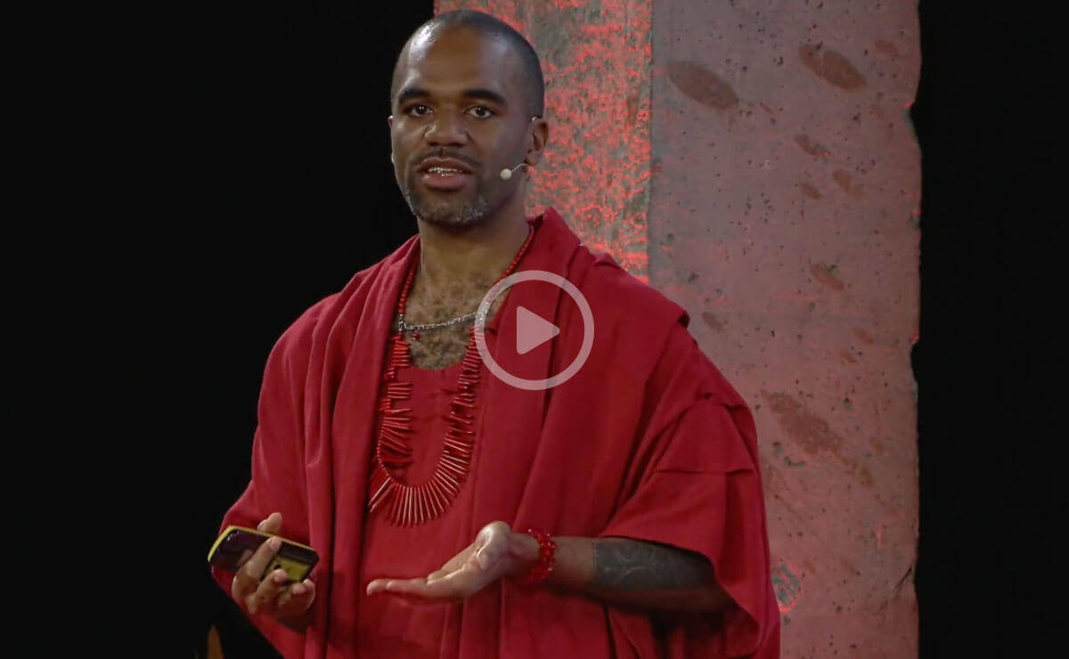 A Black man with a shaved head and wearing a red robe stands in front of a concrete pylon. There is a microphone hooked over his left ear with the end of the mic near his mouth. He holds a presentation remote control device in his right hand. He appears to be addressing an unseen audience.