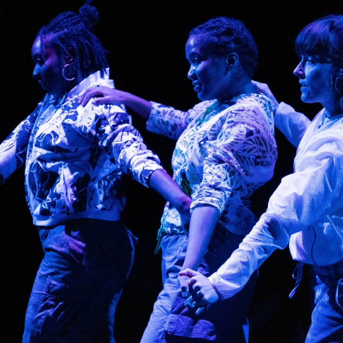 Three young women — two Black and one white — stand in a line with their right hands on the shoulder of the woman in front of them. Their left hands extend out and appear to touch. They are wearing patterned light-colored jackets and dark cargo pants. Intense blue and purple lighting on the otherwise dark stage illuminates the performers.