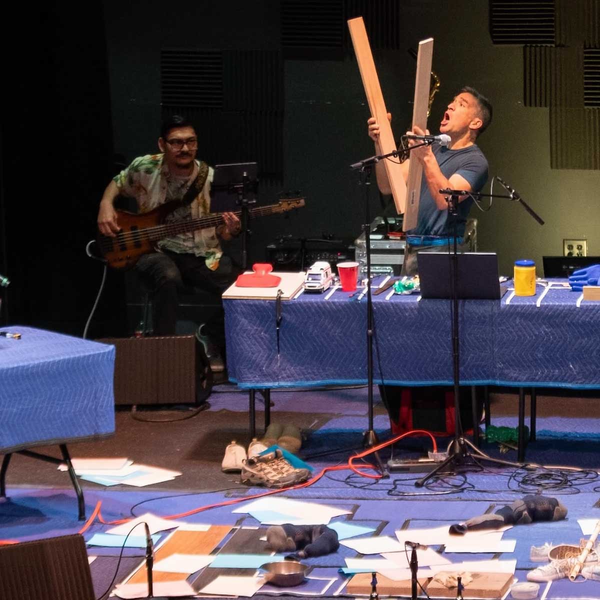 A chaotic performance space with papers and items of clothing strewn across the floor alongside long electrical cables. On the right side of the image, a man wearing a dark short-sleeved t-shirt stands behind a long rectangular table draped in blue tarp and covered with an assortment of household items, including what looks like a red plastic cup, a toy truck, and a plastic container with a lid. A laptop is also open on the table, and there are two microphone stands in front of the table and pointed at the man. He is holding in each hand a  ¼” by 4” wood board that is approximately 3 feet long, and he appears to be in the act of slapping the boards together like cymbals. His head is tilted up and his mouth open as he sings. Softly illuminated behind him in the upper left quadrant of the image, a musician wearing a green, yellow, and white shirt and dark-framed glasses sits playing a guitar. 