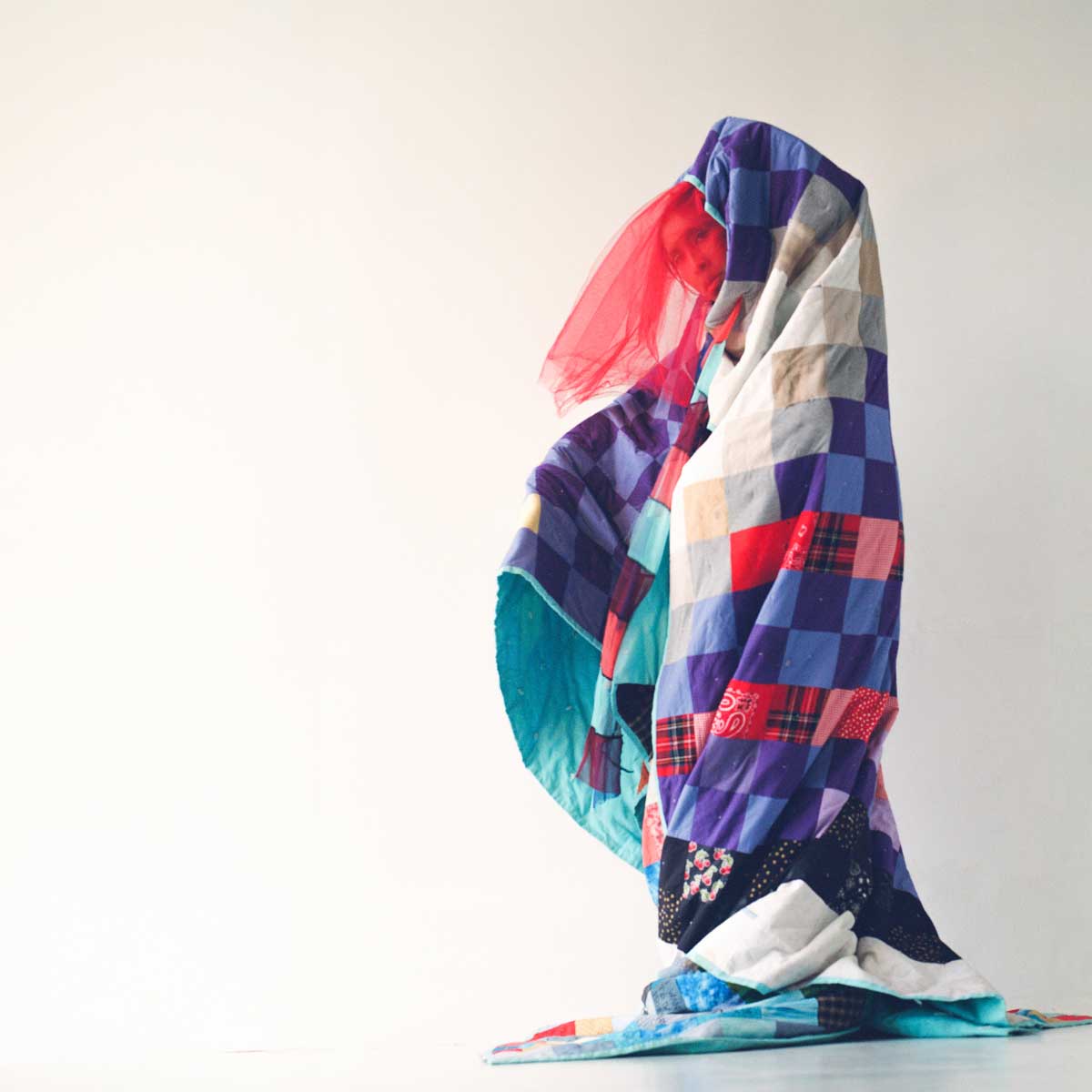 Against a blank white wall, a performer stands fully draped in a handmaid quilt consisting of a grid of squares in white, beige, multiple shades of blue, multiple shades of red, and red tartan. The inner lining of the quilt is a light teal. The performer’s head is covered by the blanket, but their face is partially visible behind a long red tulle veil.