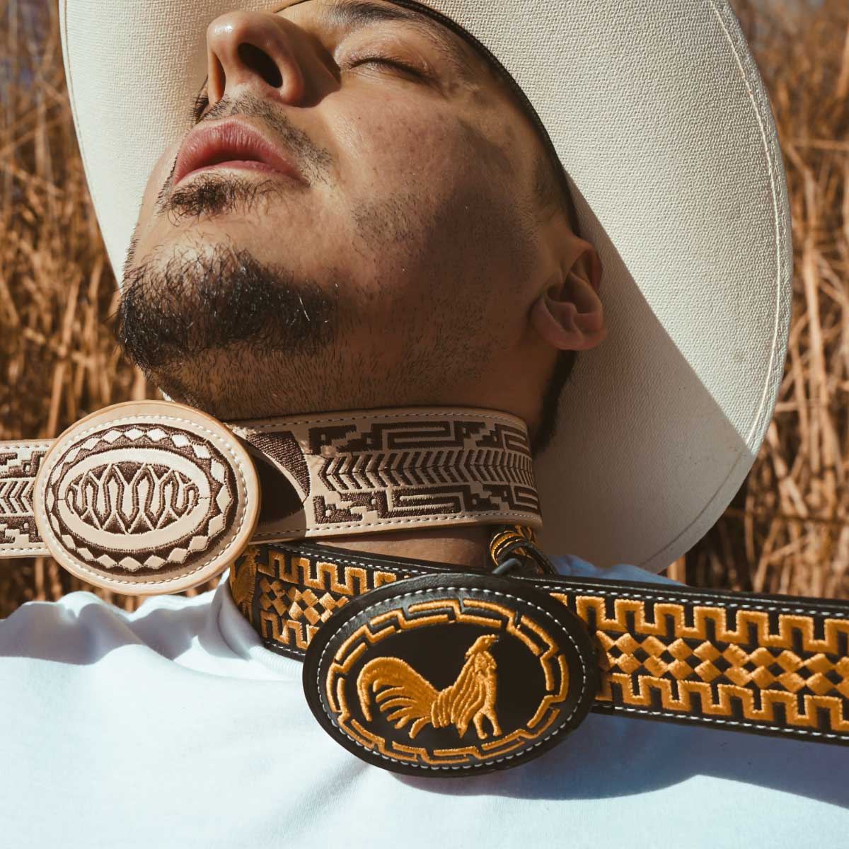 A brown-skinned young man with a slight black mustache and goatee, and wearing a pale blue t-shirt and a white cowboy hat, is leaning back with his head tilted up and eyes closed, as if he is unconscious. Two leather belts, one in tan and brown and the other in black and gold, and each decorated with what appear to be Indigenous design patterns, are wrapped around his neck, with the belts extending out of frame on either side of the image. Both belts have decorated leather oval buckles: the tan and brown buckle is decorated in geometric shapes that complement the pattern on the belt, and the black and gold buckle features a rooster surrounded by a simple border made of nesting zig-zag shapes. In the background, slightly out of focus, is yellow hay.