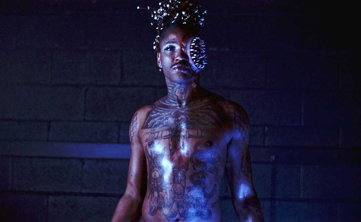 Performer Juicebox P. Burton, visible from the waist up, stands in a dark space, their body illuminated on one side by a cool white light and on the other side by a blue light, and stares intently into the camera. They are unclothed, and tattoos cover their torso, chest, shoulders, and neck. They have blue and silver eye shadow, and the left side of their face has been transformed with prosthetics and makeup into a large, nightmarish circular mouth filled with concentric rows of sharp teeth. Their hair is decorated with what appear to be small silver or white spheres that reflect the light.