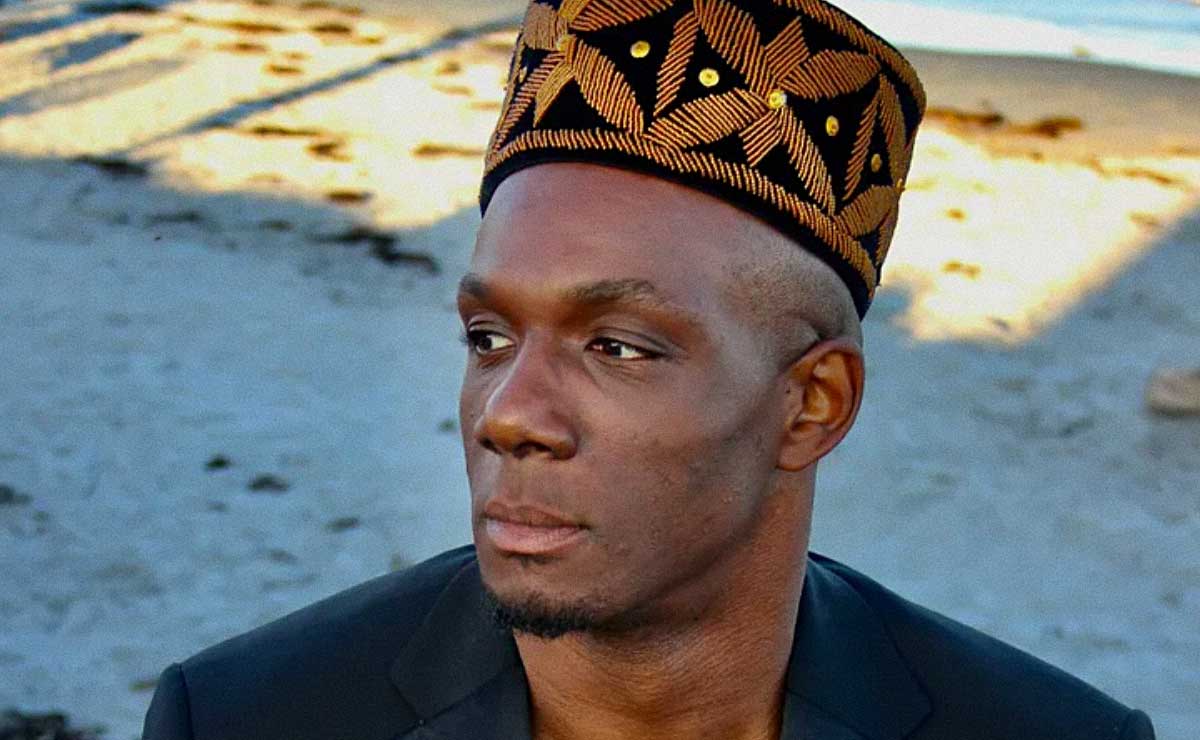 A Black man, Harold Steward, stands in shade on a beach and looks off-camera to his right, his face in three-quarter profile. The camera angle is slightly higher than his eye line. His head is shaved and he has a modest, closely cropped goatee. He is wearing a brimless, cylindrical kofia hat that's black with a repeating pattern embroidered in gold, and a black blazer or suit jacket. Behind him and out of focus is a beach, with just a hint of light blue ocean in the upper right corner of the frame. The photo appears to have been taken either early in the morning or near sunset because most of the beach is in shadow.