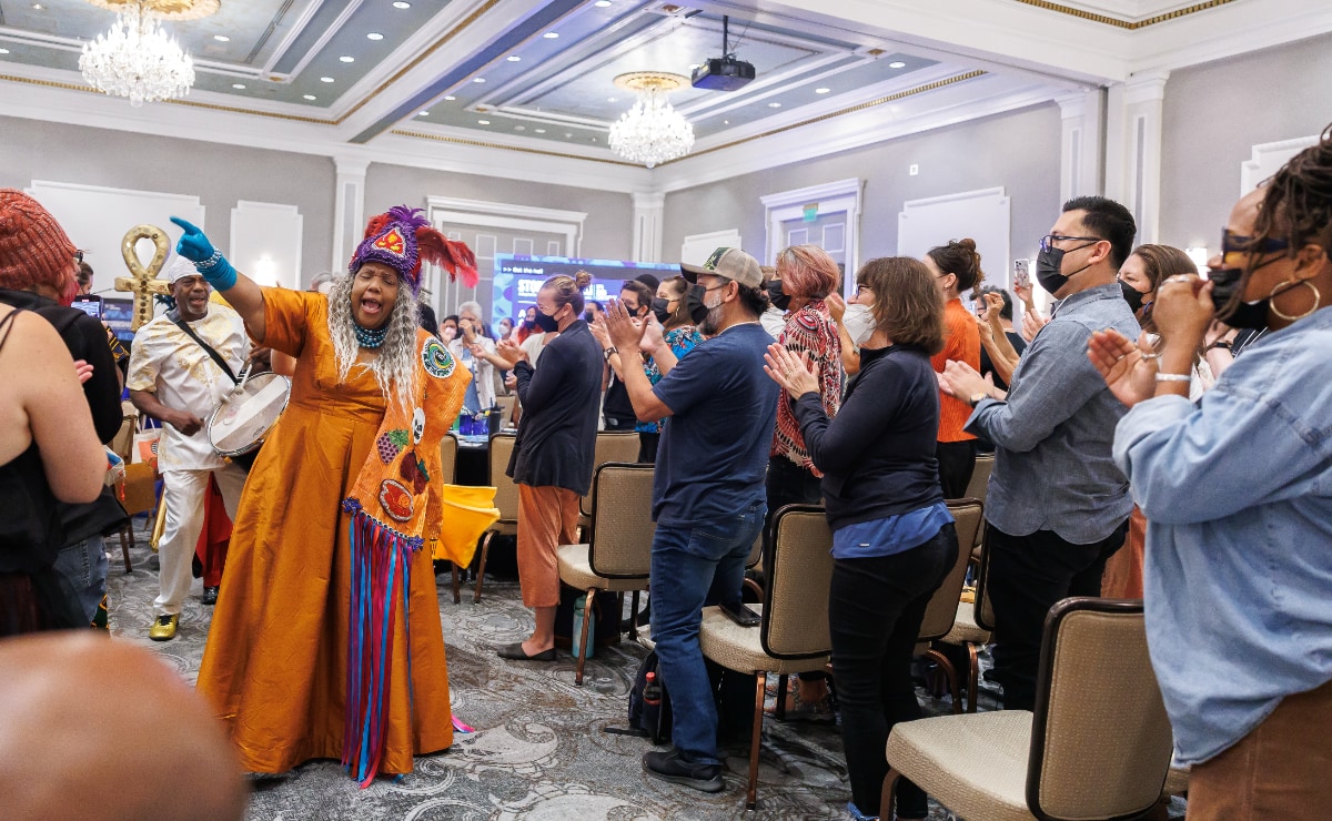 In a large, well-lit hotel ballroom with gray walls, a Black woman wearing a floor-length marigold colored dress, turquoise gloves, and a purple and red feathered headdress leads a processional through an audience of conference attendees. The attendees, all of whom are wearing face masks, are standing and clapping. The woman is singing and pointing into the air with her right hand. Behind her, a Black man wearing a white suit with gold embroidered filigree embellishments and gold shoes is accompanying her with a drum, and behind him someone is holding a large gold Egyptian ankh symbol. Draped over the woman's left shoulder is a wide sash made of the same material as the dress, with large colorful patches that include representations of a skull, a bunch of purple grapes, and two eggplants. Long blue and red ribbons hang from the end of the sash to the carpeted floor of the ballroom.