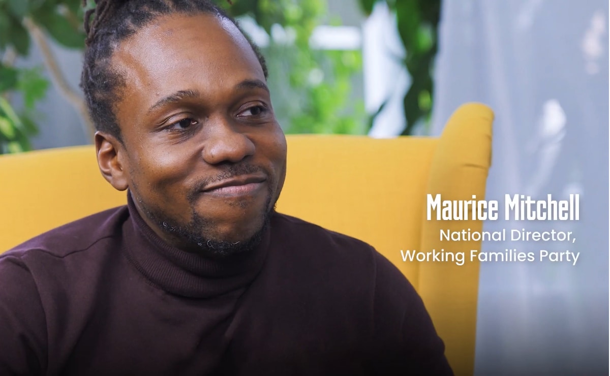 Film still of a young Black man with a neatly trimmed short beard, wearing an aubergine turtleneck and sitting in a yellow winged-back chair. He is smiling gently as he listens to an offscreen participant. White text appears to his right that reads, “Maurice Mitchell, National Director, Working Families Party.”