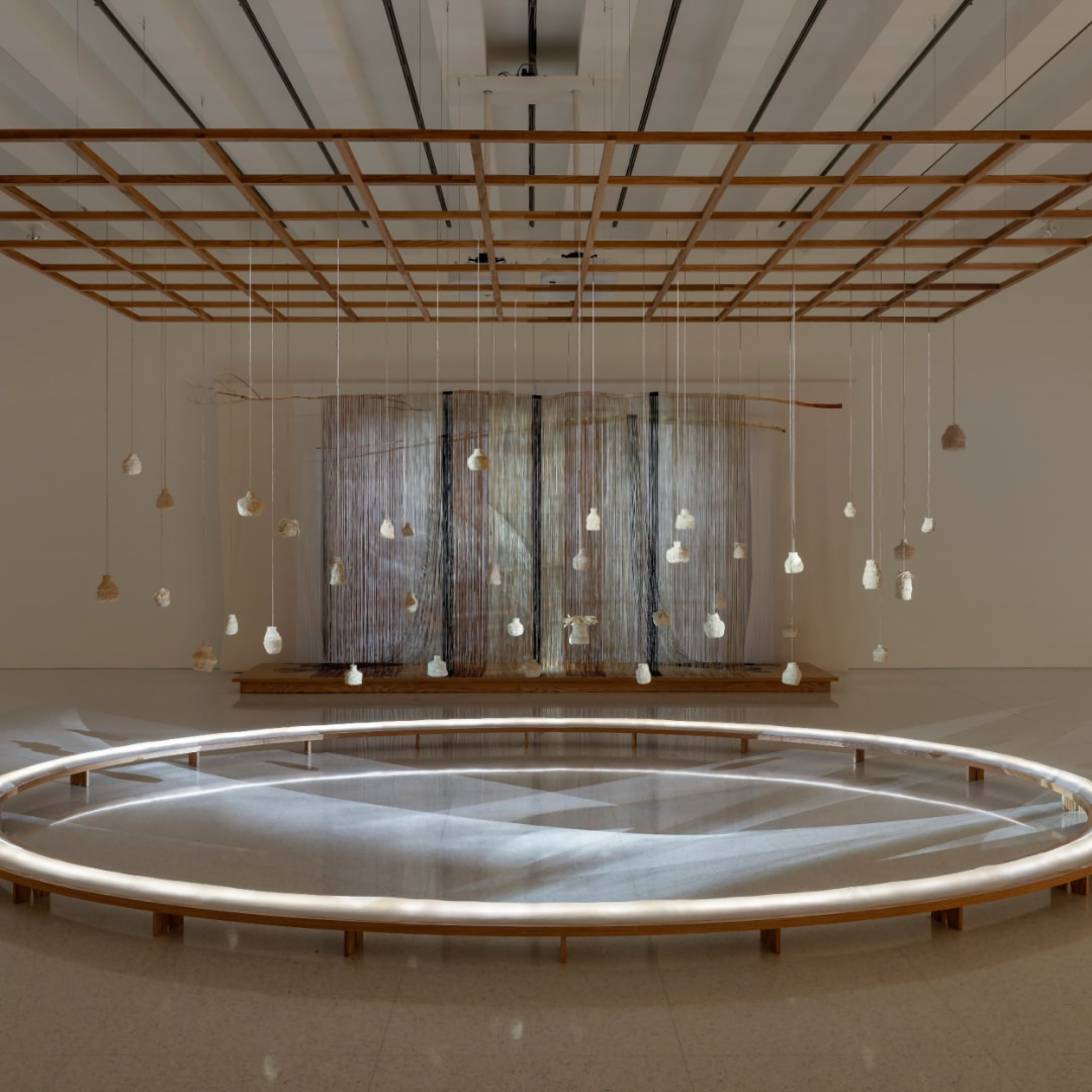 A large circular light installation on the floor of a gallery space is surrounded by suspended stones and a wooden grid overhead, with sheer curtains and subtle patterned wall panels in the background.