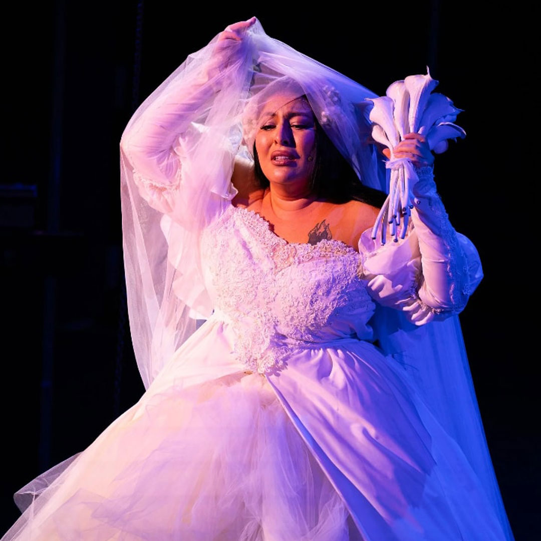 A Mexican-American woman in a white wedding dress holds her veil above her head with her right hand to reveal her face, and with her left hand holds up a simple bouquet of entirely white Calla lilies. She looks down, lips slightly parted, with an expression of anguish or intense emotion. Over her left breast, the top part of a tattoo is visible above the bodice of the dress. The tattoo appears to be three church steeples, each topped with a cross, surrounded by three large candle flames. The woman stands alone in a dark space with her dress and face illuminated by soft pinkish-white light, and her side illuminated by a soft blue that suggests moonlight.