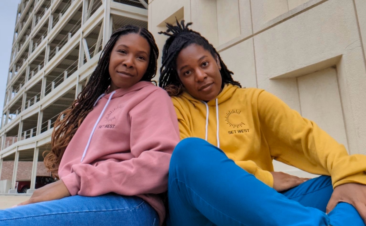 Two young Black women in jeans and sweatshirts sit outside in a city environment and smile gently at the camera. Partially obscured in the background is a large sand colored building without windows, and an adjoining sand colored multi-story parking garage. On the left, Chasah West, wearing a muted pink sweatshirt and blue jeans, sits with her back against her sister's shoulder and her face turned toward the camera. She is smiling gently. On the right, Charliese West, wearing a gold colored sweatshirt and blue jeans, sits cross-legged and stares at the camera with a neutral but friendly expression.