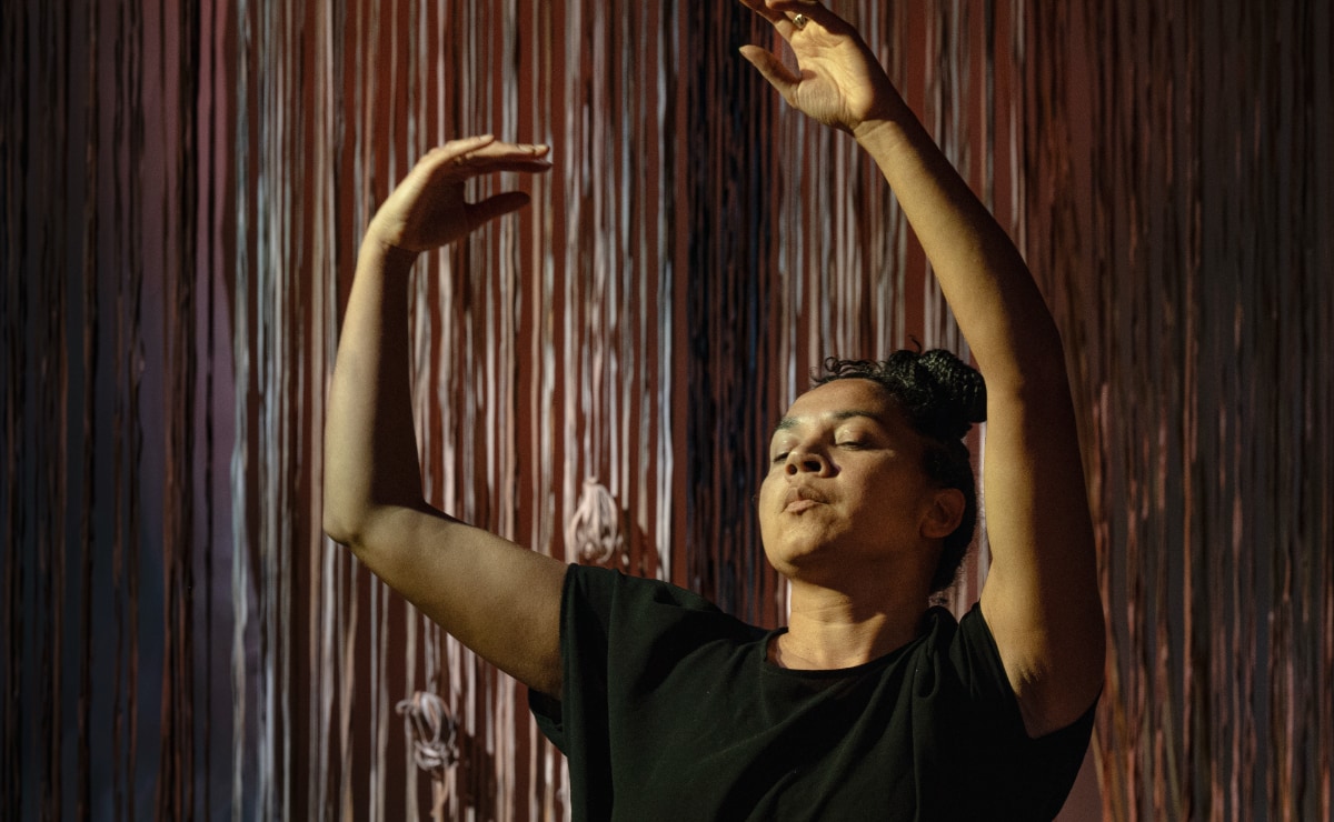 A Black, female-presenting dancer in a black short-sleeved shirt is shown from the mid-torso up with her head tilted back slightly and her eyes closed. She extends her left arm above her head in a gentle curve, and holds her right arm in a bent position above shoulder level. She is warmly illuminated in what appears to be an otherwise dark performance space. The backdrop behind her is covered in narrow, russet-colored vertical lines, with curls of dried leaves near the bottom, suggesting an abstract representation of plant stalks, plant fiber, and twine.