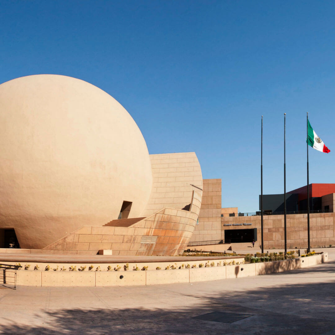 Exterior shot of the distinctive spherical IMAX theater building on the grounds of the Tijuana Cultural Center in Tijuana, Mexico. The windowless, smooth sphere is approximately five stories tall and is made of concrete tinted a sandy light brown. The photo is cropped to show only the middle and right half of the sphere. A curving terraced staircase that wraps around the side of the sphere suggests a cutaway view of a nest or protective eggshell. Three flagpoles stand to the right of the building, and Mexico's national flag waves from the outermost pole. The sky is blue and free of clouds. In the background is a partial view of the Cultural Center’s other more conventional buildings.