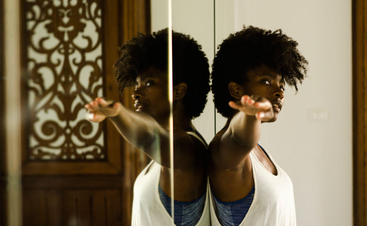 Melanie Greene stands with her back to a mirrored wall and extends her right arm toward the camera as she looks into the camera lens. Her shoulder is barely touching the mirror, and her reflection is undistorted and in focus, creating a symmetrical twinning effect between Melanie and her reflection. She wears a loose fitting plain white tank top layered over a heather gray tank top. The mirror also reflects a brown wooden door somewhere in the background, with a narrow window protected by an intricately carved decorative wood screen. 